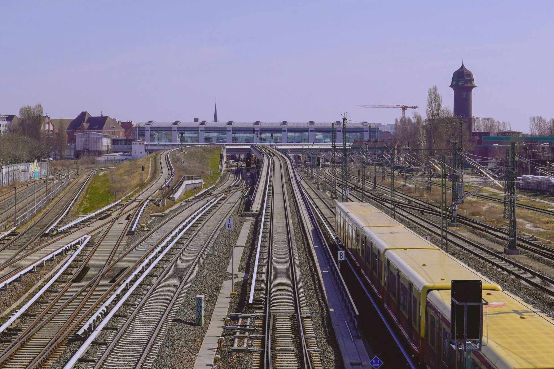 Mehrgleisiger Bahnknoten mit Oberleitungen und gelbem Zug, Blick auf Weichen und Bahnhofsbauten.