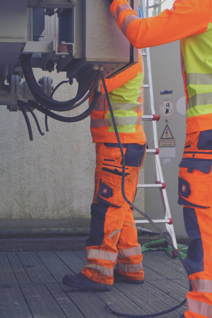 Zwei Personen in Warnkleidung arbeiten an einem geöffneten Schaltschrank neben einer Leiter; Kabel hängen herunter, Warnschild „Hochspannung“.