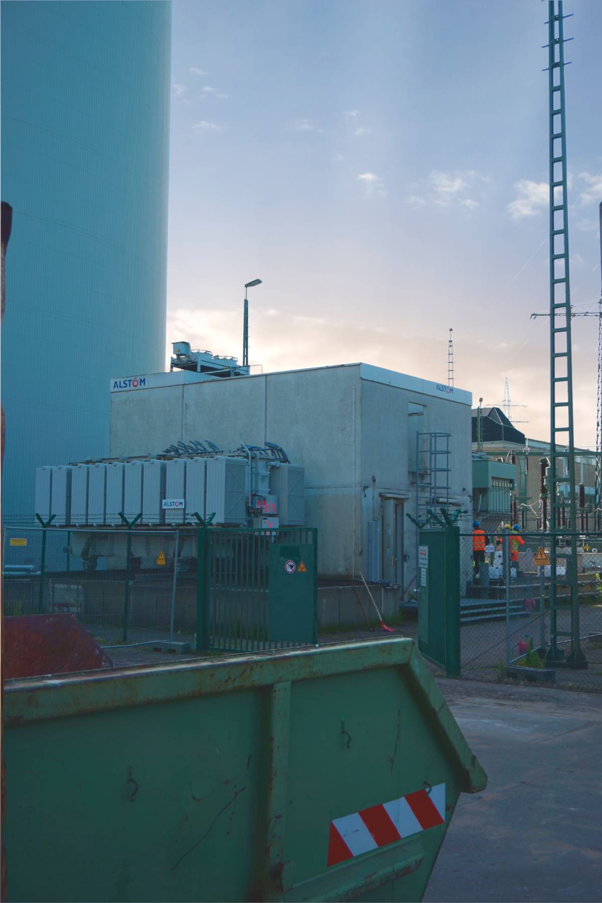 Transformatorstation auf einem Industriegelände, umzäuntes Areal mit Container im Vordergrund bei Tageslicht.