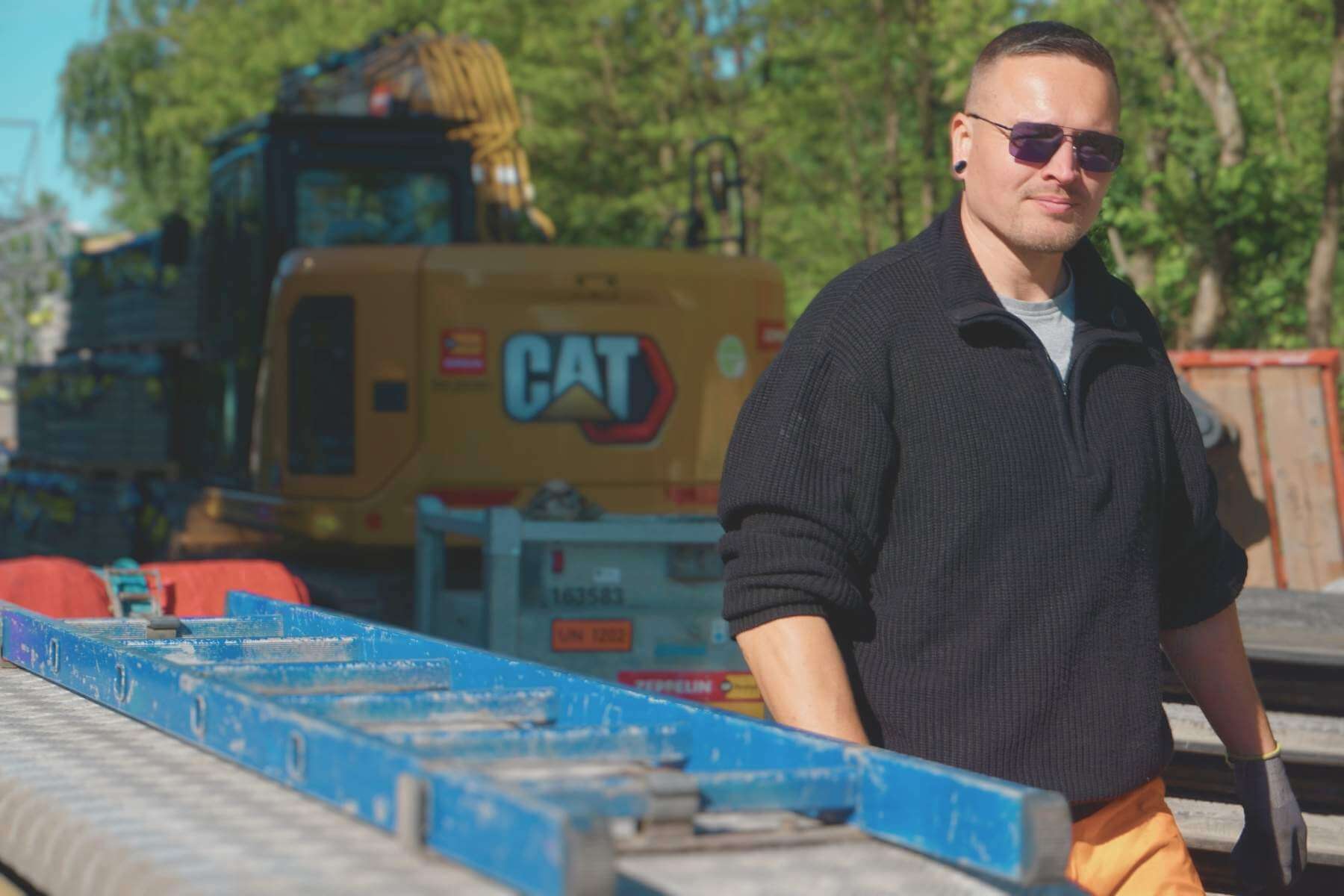 Mitarbeiter mit Schutzbrille und Handschuhen neben Tieflader; unscharfer Bagger im Hintergrund