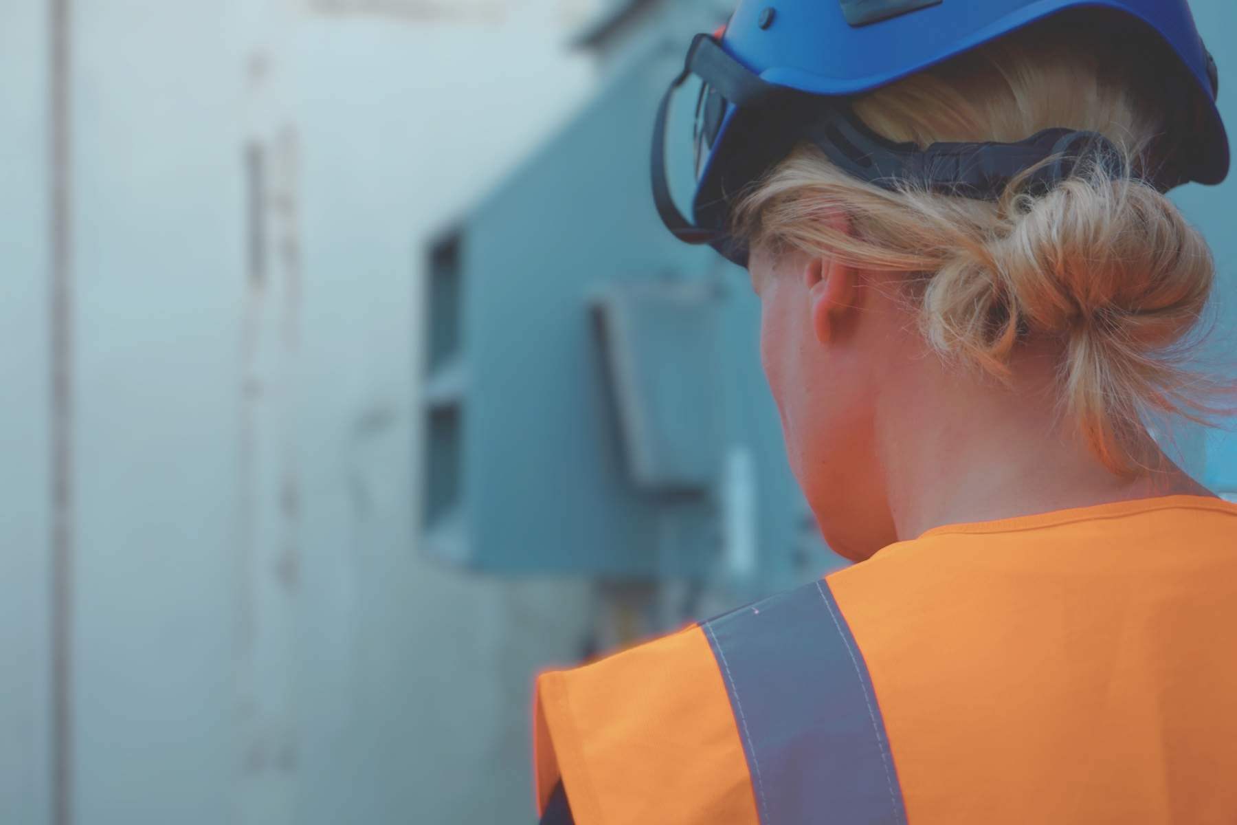 Person mit Schutzhelm und Warnweste steht vor einem Schaltschrank