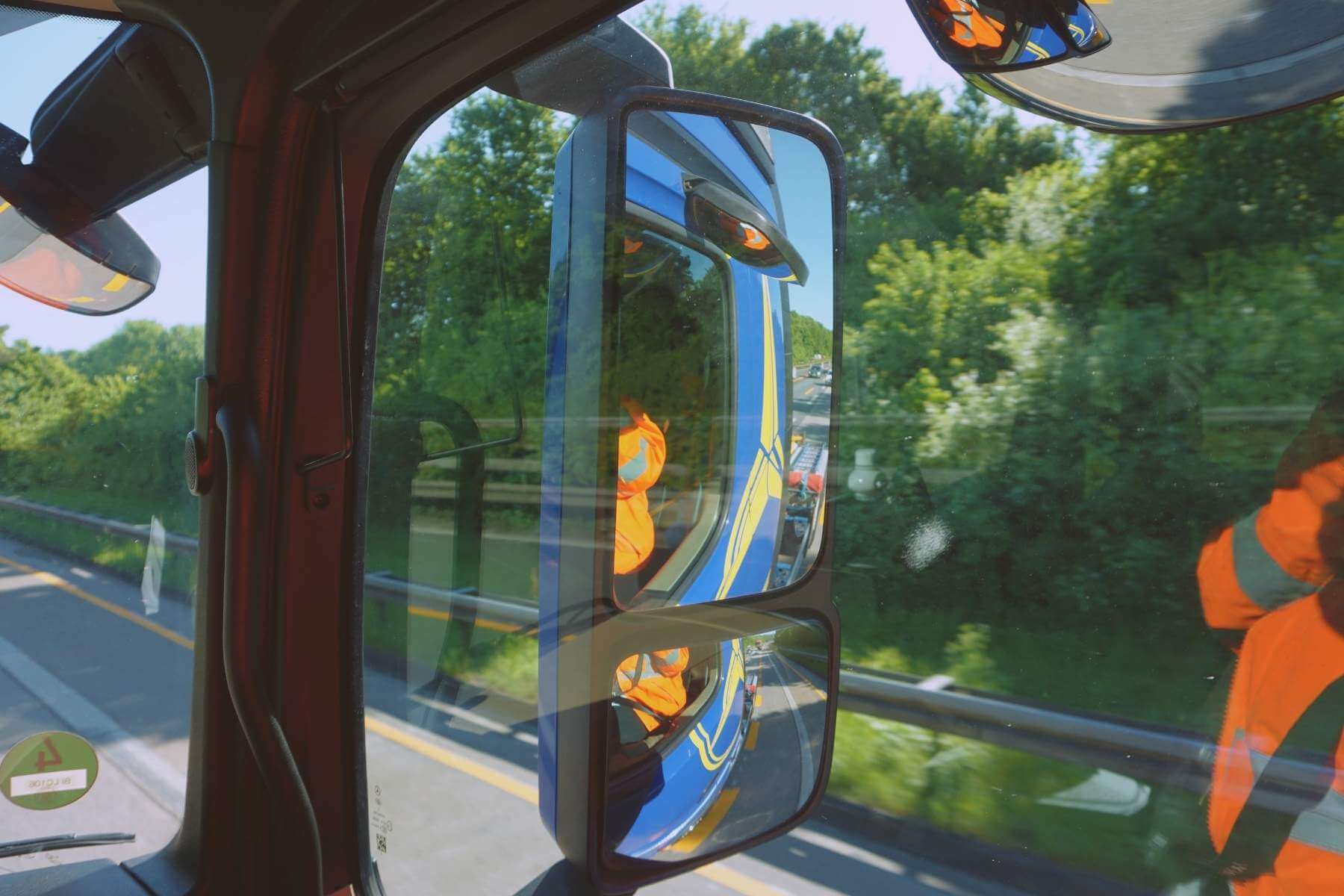 Blick durch LKW-Seitenfenster auf Außenspiegel; Reflexkleidung spiegelt sich, Autobahn im Hintergrund