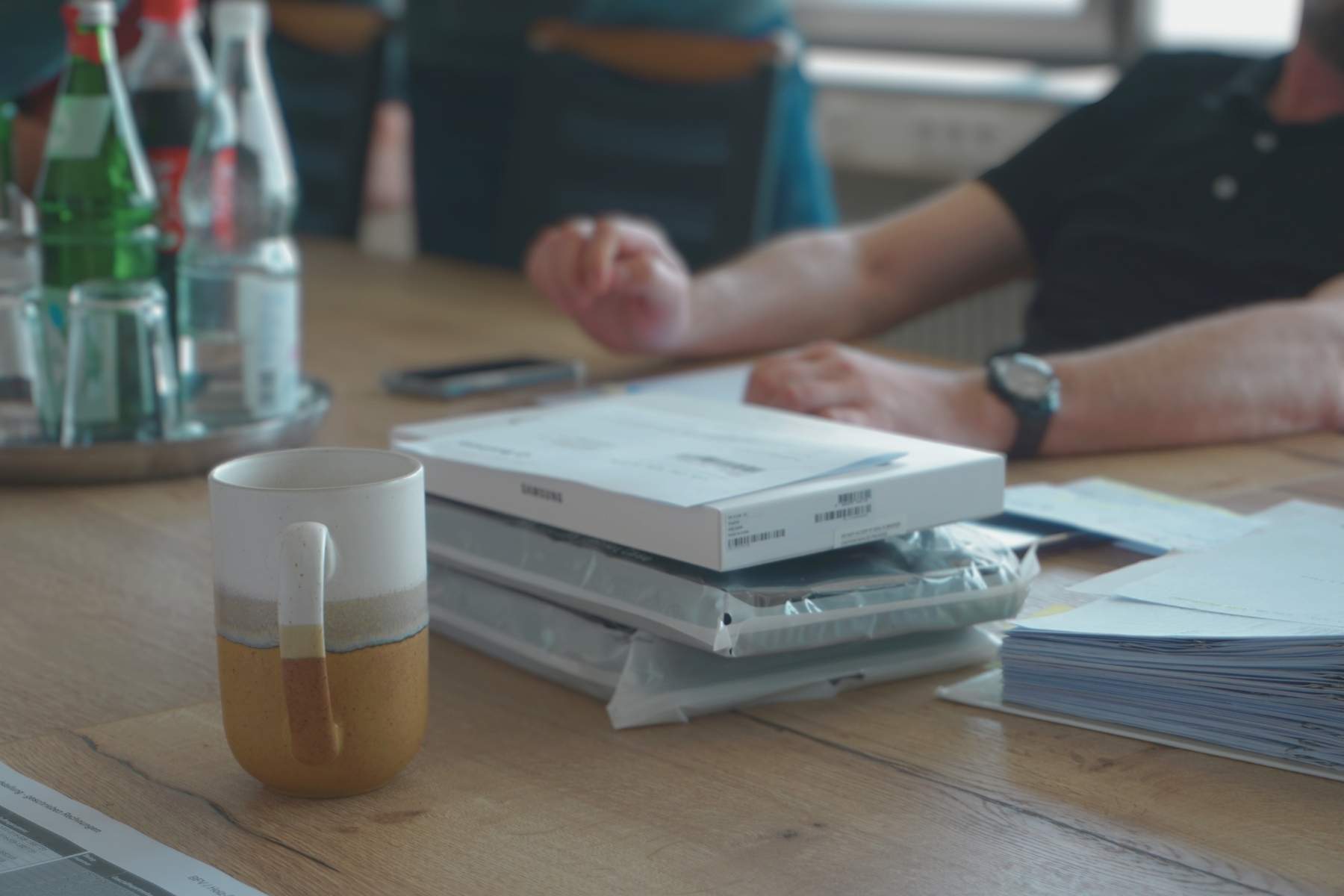 Meetingtisch mit Kaffeebecher, Wasserflaschen, Dokumentenstapeln und verpackten Geraeten; Personen ohne erkennbare Gesichter im Hintergrund.