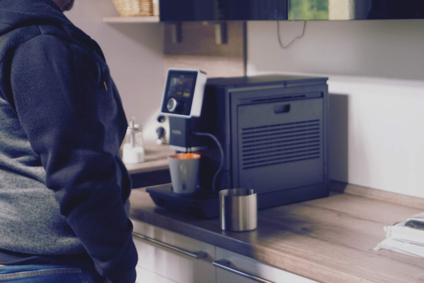 Person steht an einer Kaffeemaschine in der Büroküche und bereitet sich ein Heißgetränk zu.
