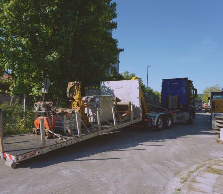 Tieflader mit geladenen Anbauteilen an der Laderampe auf einer Baustellenzufahrt; Zugmaschine im Hintergrund